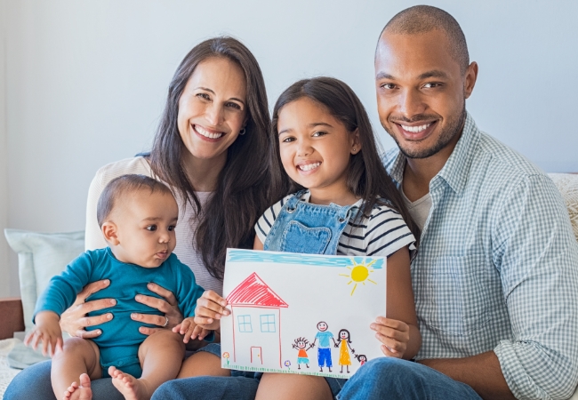 Multicultural family, mom, dad, daughter, and baby boy, sitting on couch. Daughter is holding a drawing of family by their home.